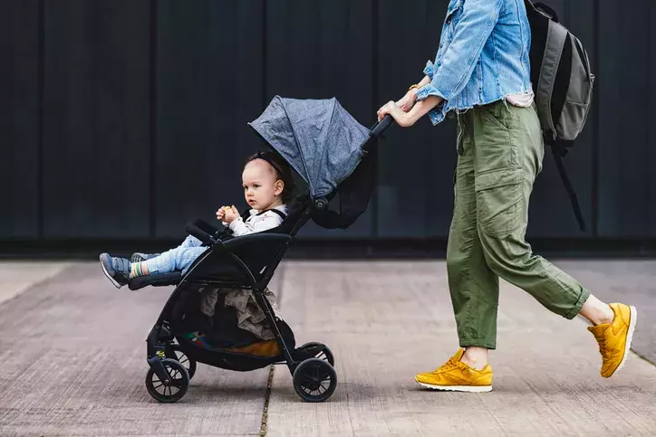 Femme en pantalon vert et baskets jaunes poussant une poussette avec un bébé sur un trottoir